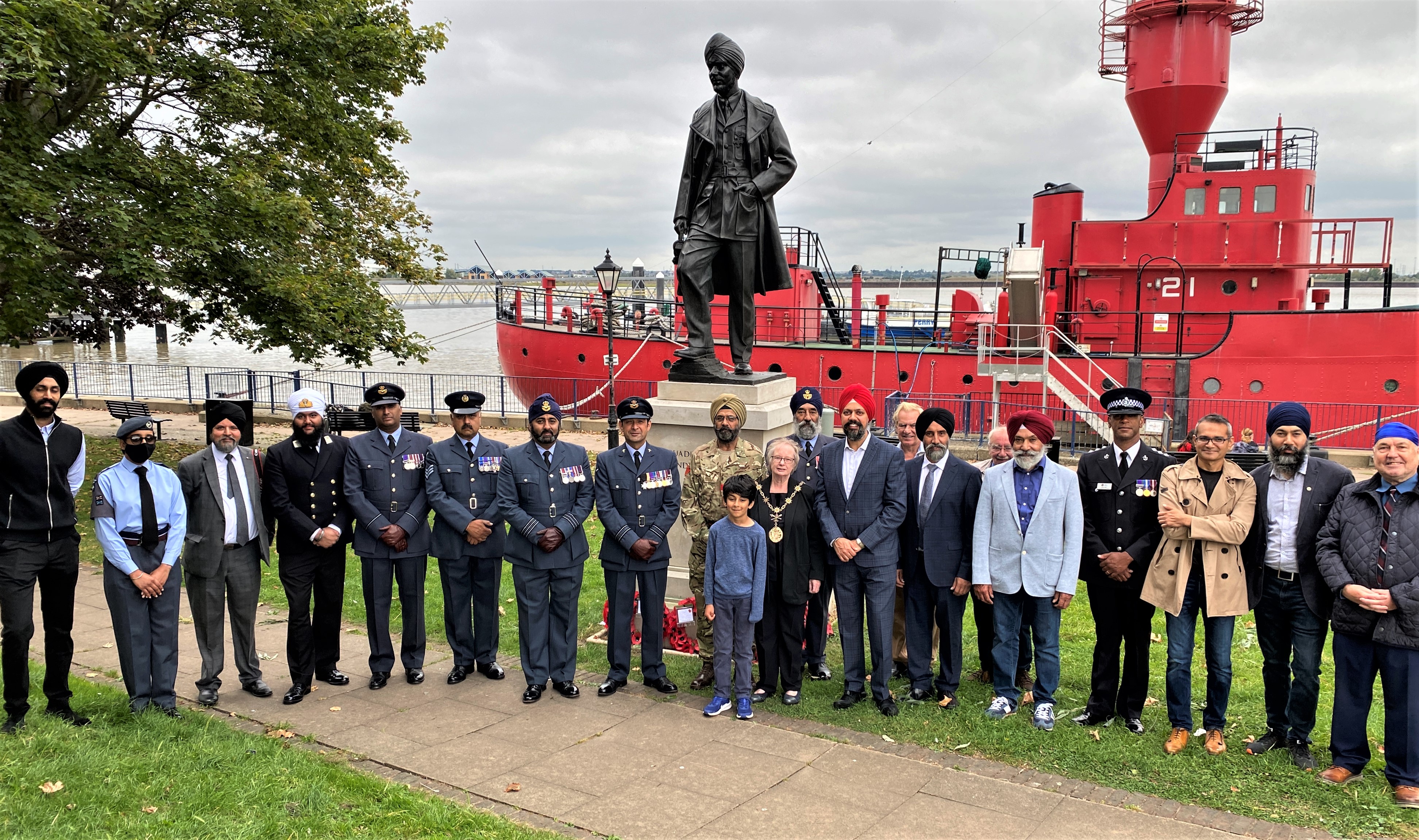 Personnel and family gather next to statue, with boat on the river in the background.